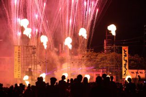 花火が背景に上がるステージとそれを見る人々の夜景の写真